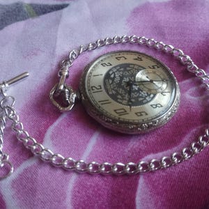 May include: A silver pocket watch with a chain, resting on a pink floral fabric. The watch face is cream-colored with black numerals and a smaller subdial. The chain has a T-bar clasp.