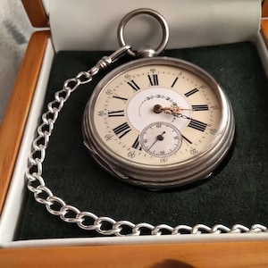 May include: A silver pocket watch with a white face and black Roman numeral hour markers. The watch has a small second hand dial and is in a wooden box with a green velvet lining.