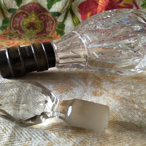 May include: Two clear glass bottle stoppers. One is a round, faceted stopper with a metal cap. The other is a faceted, diamond-shaped stopper with a white top.