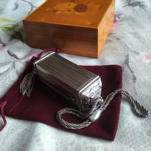 May include: A silver box with an ornate design and a tassel. The box is sitting on a burgundy velvet pouch with a drawstring.