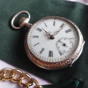 May include: A silver and gold pocket watch with a white face and black Roman numerals. The watch has a small seconds dial and ornate detailing around the bezel. It rests on a dark green velvet pouch, with a gold chain in the foreground.