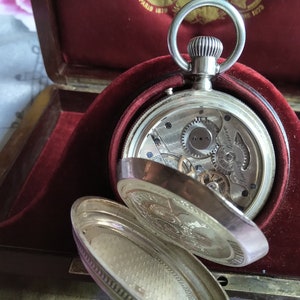 May include: A silver pocket watch with an open face, revealing the intricate clockwork mechanism. The watch is resting in a red velvet-lined box.