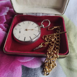 May include: A silver-toned pocket watch with a white face, black Roman numerals, and a small sub-dial, accompanied by a gold-toned chain and a winding key. The set is presented inside a rectangular case with a vibrant red velvet interior lining.