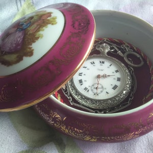 May include: A vintage pocket watch with a silver chain inside a porcelain trinket box. The box is decorated with gold trim and a painted scene of a couple in a garden.