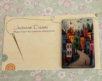 May include: A rectangular pendant with a colourful cityscape design, featuring a winding road, trees, and buildings in blue, red, orange, and yellow. The pendant is on a card with the text "Inchworm Designs" and "Make room for creative adventures."