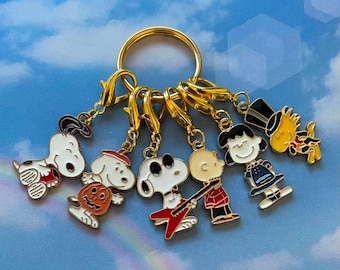 May include: A gold-toned metal keyring featuring six dangling enamel charms depicting Peanuts characters. The charms include Snoopy, Snoopy with a pumpkin, Snoopy with sunglasses and a red guitar, Charlie Brown, Lucy, and Woodstock wearing a top hat.