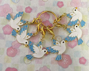 May include: A collection of white bird charms with blue wing and tail accents, attached to gold-coloured clasps and a ring. The charms are arranged in a circular pattern, set against a floral patterned background.