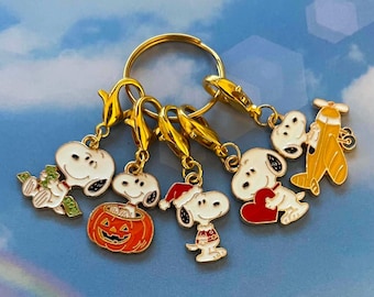 May include: A gold-toned metal keyring featuring five enamel charms of a white beagle character. The charms depict the character holding green money, sitting in an orange pumpkin, wearing a red Santa hat, holding a red heart, and flying a yellow plane.
