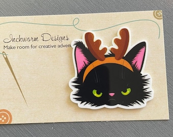 May include: A black cat sticker with a grumpy expression, wearing a brown antler headband and pink-lined ears. The sticker is on a tan card with the text "Inchworm Designs" and "Make room for creative adventures."