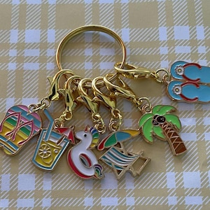 May include: A gold keyring with various charms, including colourful flip-flops, a tropical drink, a unicorn float, a beach chair, a palm tree, and blue flip-flops. The charms are attached with gold clasps.