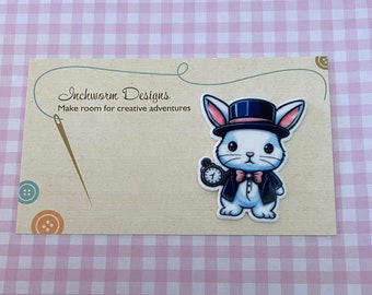 May include: White rabbit wearing a black top hat, blue jacket, and bow tie, holding a pocket watch with a Roman numeral one. The rabbit is a cartoon illustration on a white background. The text "Inchworm Designs" and "Make room for creative adventures" is printed on a beige background.
