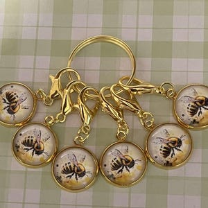 Puede incluir: Conjunto de seis marcadores de punto de abeja dorados con un anillo dorado. Cada marcador presenta un encanto redondo de vidrio con un diseño de abeja pintado.