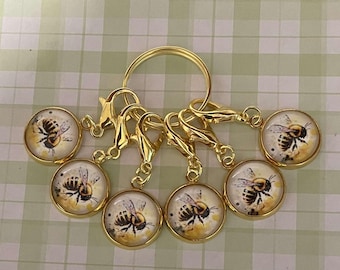 May include: Set of six gold bee stitch markers with a gold ring. Each marker features a round glass charm with a painted bee design.