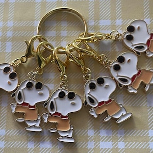 May include: A set of Snoopy keyrings. Each keyring features a white Snoopy figure with black eyes, a red collar, and a tan shirt. The keyrings are attached to gold-coloured clasps and a large ring.