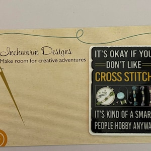 Op de afbeelding: Een zwarte en gele sticker met de tekst "It's okay if you don't like cross stitch. It's kind of a smart people hobby anyway." De sticker toont een kruissteekring, een bol garen en borduurdraad.