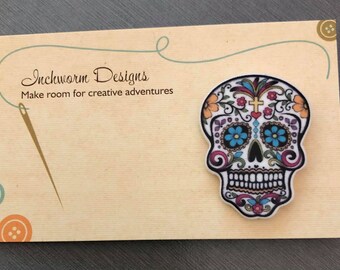 May include: A decorative sugar skull badge with a vibrant design, featuring flowers and a cross. The badge is displayed on a tan card with the text "Inchworm Designs" and "Make room for creative adventures".