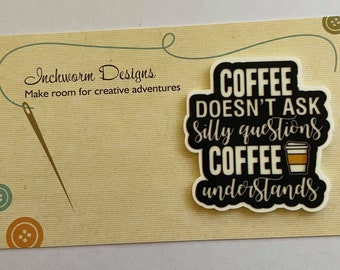 May include: Black and white sticker with the text "Coffee doesn't ask silly questions, Coffee understands" and a cartoon image of a coffee cup. The sticker is on a beige background with a needle and thread and a few buttons.