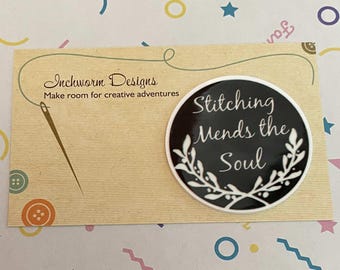 May include: A black and white round pin with the text "Stitching Mends the Soul" surrounded by a white floral design. The pin is on a brown card with the text "Inchworm Designs Make room for creative adventures".