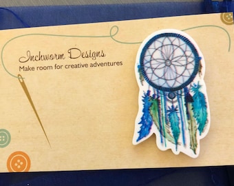 May include: A blue and green dreamcatcher with feathers hanging from the bottom. The dreamcatcher is on a brown card with the text "Inchworm Designs Make room for creative adventures".