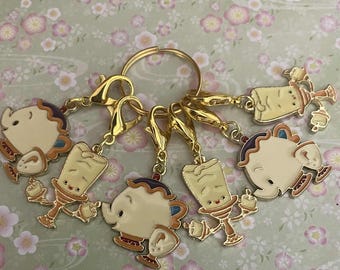 May include: A gold-toned keychain featuring six enamel charms. The charms depict characters from a fairy tale, including a teapot, a teacup, and a candlestick. The charms are attached to a gold ring with lobster claw clasps.
