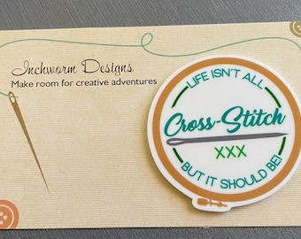 May include: A brown and white circular sticker with the text "LIFE ISN'T ALL CROSS-STITCH BUT IT SHOULD BE" and a needle and thread design. The text "XXX" is in green.