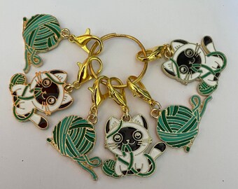 May include: A set of gold-coloured metal stitch markers. Each marker features a white and black cat design with green yarn balls. The cat charms have gold accents and are attached to gold-coloured lobster clasps and a gold ring.