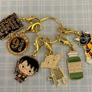 May include: A set of six gold-coloured Harry Potter themed charms. The charms include a round charm with the text "Platform 9 3/4", a charm with the text "I solemnly swear that I am up to no good", a charm with a cartoon image of Harry Potter, a charm with a cartoon image of Dobby the house elf, a charm with a cartoon image of a black cat wearing a Hogwarts scarf, and a charm with a green bottle that says "Felix Felicis Potions".