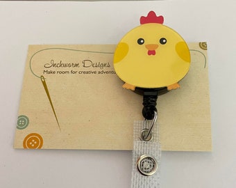 May include: A yellow chick keychain with a red comb and black eyes. The keychain is attached to a black retractable badge holder with a white strap. The background is a tan card with the text "Inchworm Designs Make room for creative adventures".