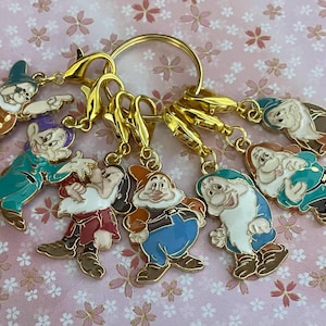 Könnte beinhalten: Eine Sammlung von sieben farbenfrohen Emaille-Charm-Schlüsselanhängern mit den sieben Zwergen aus Schneewittchen. Jeder Anhänger ist mit goldfarbenen Metallakzenten versehen und an einem goldenen Schlüsselring befestigt. Der Hintergrund ist ein rosa Blumenmuster.