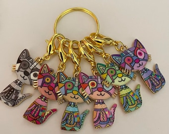 May include: A set of six colourful cat charms with gold-tone metal rings. The charms are attached to a gold-tone metal key ring. The cats are all different colours and have different patterns.
