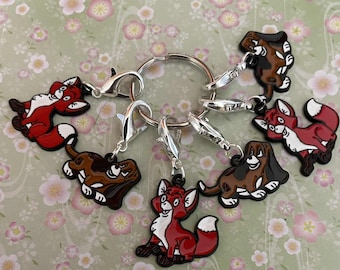 May include: A collection of cartoon fox and dog character charms in red, white, and brown, attached to silver-colored lobster clasps and a key ring. The charms are arranged on a floral patterned background.