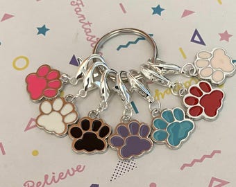 May include: A set of seven silver-toned stitch markers with paw print charms in various colours, including pink, white, black, purple, teal, and red. The charms are attached to a silver key ring.