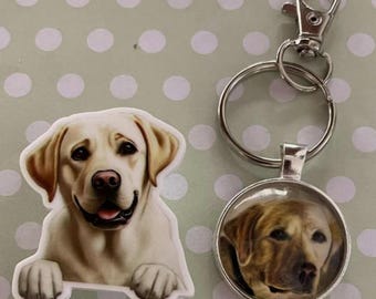 May include: A sticker and a keyring featuring a Labrador Retriever. The sticker shows a smiling yellow Lab. The keyring has a silver-coloured ring and clasp, with a round pendant showing a Lab's face.
