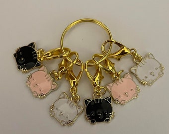 May include: A collection of gold-coloured metal cat-shaped charms with lobster clasps, attached to a gold ring. The charms are in black, white, and pink enamel. The cat charms have black details and are arranged in a circular pattern.