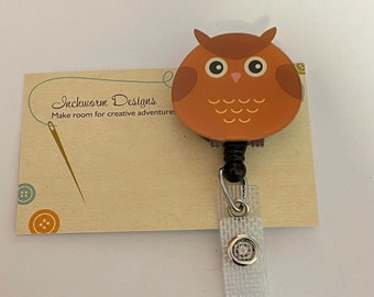 May include: A brown owl-shaped retractable badge holder with a white strap and a silver button. The badge holder has a black cord and is attached to a card with the text "Inchworm Designs Make room for creative adventures."