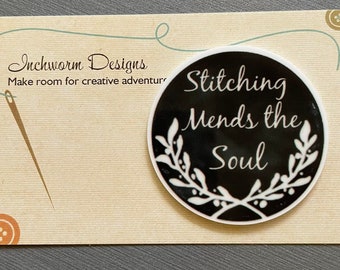 May include: Black and white round sticker with the text "Stitching Mends the Soul" surrounded by a white floral design. The sticker is on a beige background with the text "Inchworm Designs Make room for creative adventures".