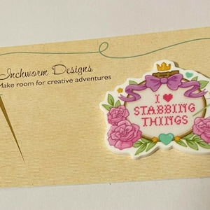 May include: A white, pink and green embroidery hoop with a pink bow and a crown on top. The hoop has a cross-stitch design that says "I love stabbing things". The hoop is on a beige card with the text "Inchworm Designs Make room for creative adventures".