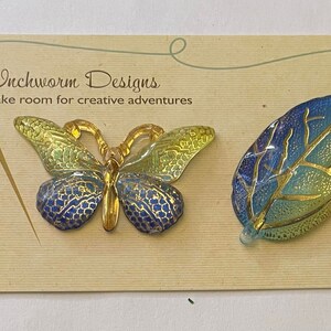May include: A butterfly and leaf-shaped decorative items. The butterfly has gold, blue, and yellow accents. The leaf is blue and yellow with gold veins. The items are on a card that reads "Inchworm Designs".