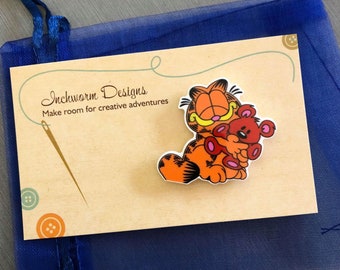 May include: A wooden pin featuring a cartoon orange cat hugging a red teddy bear. The pin is on a beige card with the text "Inchworm Designs Make room for creative adventures".