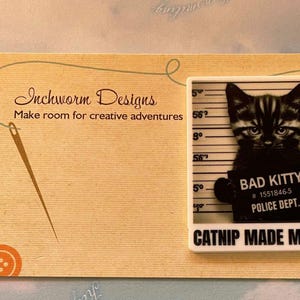 May include: A white square pin with a black and white image of a cat in a police lineup. The cat is wearing a sign that says "Bad Kitty #1551846-5 Police Dept." The text below the image says "Catnip Made Me Do It."