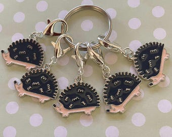 May include: Five silver keychains with black and pink enamel hedgehog charms. Each charm has a different design of a hedgehog with a pink belly and black spines.