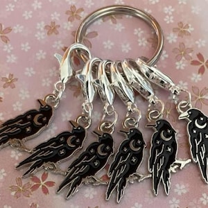 May include: A silver keyring with six black raven charms. Each raven features a crescent moon and star design. The charms are attached to a silver ring and lobster clasps. The background is a pink floral pattern.