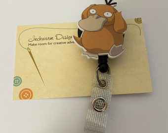 May include: A Psyduck character badge reel with a cartoon design. The badge reel is attached to a light tan card with the text "Inchworm Design" and "Make room for creative adventure". The reel has a white strap and a silver metal clip.