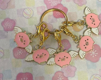 May include: A gold-coloured keyring with six pink pig charms, each with white wings. The charms are attached with gold-coloured clasps and a large ring. The background features a floral pattern.