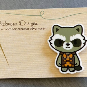 Pode incluir: Um alfinete branco de estilo de desenho animado do Rocket Raccoon dos Guardiões da Galáxia. Ele está vestindo seu macacão marrom e laranja característico. O alfinete está em um cartão que diz "Inchworm Designs" e "Make room for creative adventures".