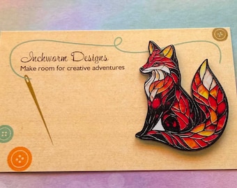 May include: A fox-shaped pin with a stained-glass style design in shades of red, orange, and yellow, mounted on a tan display card. The card features the text Inchworm Designs and Make room for creative adventures, a needle, and buttons.