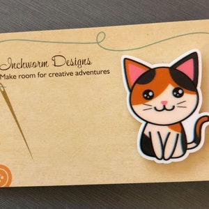 May include: A calico cat with pink ears and a white belly sits on a brown background. The cat has black eyes and a black nose. The background has the text "Inchworm Designs" and "Make room for creative adventures".