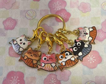 May include: A gold-colored keychain with seven cat charms. Each charm is a colorful cat design with a fish-shaped body. The cats have various colors, including orange, pink, white, and black. The background is a floral pattern.