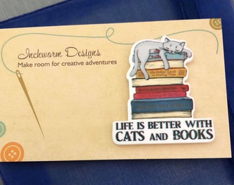 May include: A white ceramic pin with a cat sleeping on a stack of books. The pin says "Life is better with cats and books".