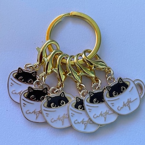 May include: A gold keychain with five charms featuring black cats peeking out of white coffee mugs. Each mug has the text "Cat Cafe" written on it.